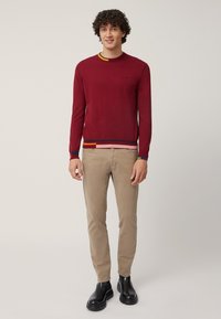 Maglione rosso lavorato a maglia con dettagli a strisce multicolori sul collo e sull'orlo, abbinato a pantaloni beige e stivaletti neri.