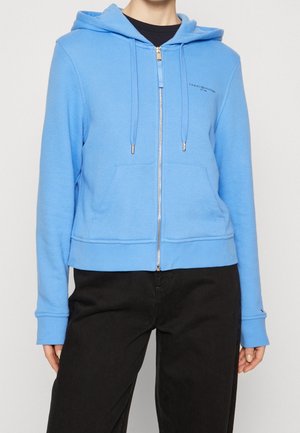 Persona que lleva una sudadera con capucha azul claro con cremallera, cordones y bolsillos delanteros, combinada con pantalones negros, sobre un fondo liso.