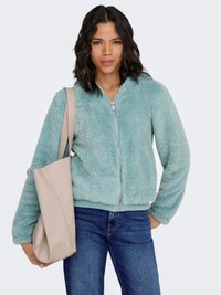 Faux fur zip-up jacket in light teal, featuring a relaxed fit and ribbed cuffs. Paired with a cream handbag and denim jeans.
