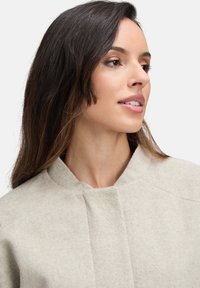 Hellgrauer, strukturierter Wollmischmantel mit hohem Kragen, der ein minimalistisches Design und dezente Fältelungen an der Vorderseite aufweist.