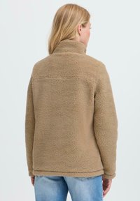 Femme aux cheveux blonds portant une veste en sherpa beige et un jean bleu, vue de dos sur un fond clair.