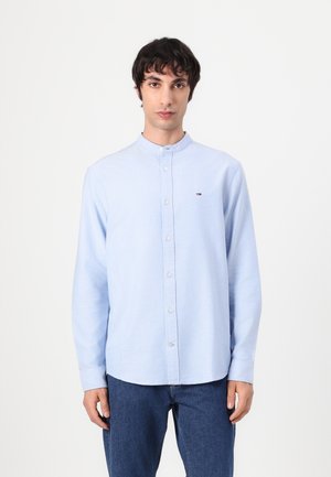 Camicia button-up azzurro chiaro a maniche lunghe con colletto alla cinese e un piccolo logo sul petto, abbinata a jeans blu scuri. Tessuto dalla consistenza liscia.