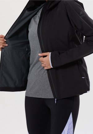 Persona que lleva una chaqueta negra sobre una camisa gris, sosteniendo la chaqueta abierta para mostrar el forro interior de malla y el bolsillo interior.