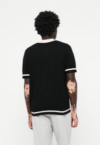 Fekete kötött pólóing fehér csíkos mandzsettával és gallérral, laza szabású és sima textúrájú. A modellen szürke nadrág van.