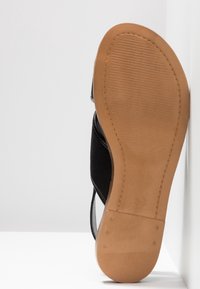 Sandale avec une semelle en caoutchouc marron clair et des brides noires, vue du dessous sur un fond blanc.