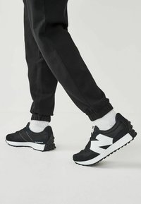 Zapatillas deportivas negras con acentos blancos, hechas de ante y malla, con una suela texturizada y un distintivo prominente en el lateral.