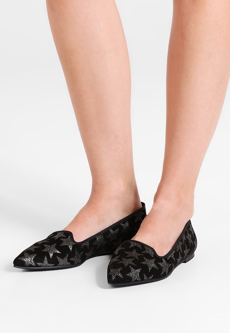 Pies con zapatos planos negros de punta con patrones de estrellas brillantes plateados sobre un fondo blanco.