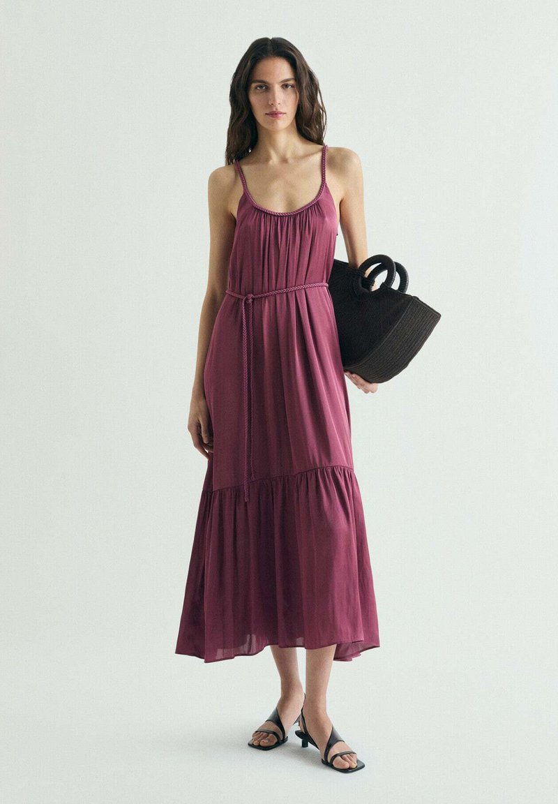 Femme portant une robe mauve sans manches à fines bretelles, tenant un grand sac à main noir tressé, et portant des sandales noires à talons.