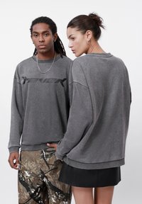 Felpe grigie con una finitura testurizzata, caratterizzate da un logo nero sul davanti. Abbinate a pantaloni in camouflage e a una gonna in pelle nera.