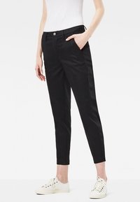 Pantaloni neri a sigaretta con una texture liscia, dotati di tasche frontali e chiusura con bottone, abbinati a sneakers sportive bianche.