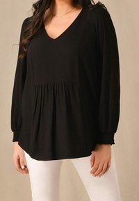 Svart blus med V-ringning, långa ärmar och smockade ärmslut. Har en texturerad tygkvalitet och en hopad midja, i kombination med vita byxor.