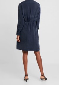 Abito blu navy realizzato in tessuto morbido, con vita arricciata, maniche lunghe e orlo fino al ginocchio. Abbinato a tacchi neri.