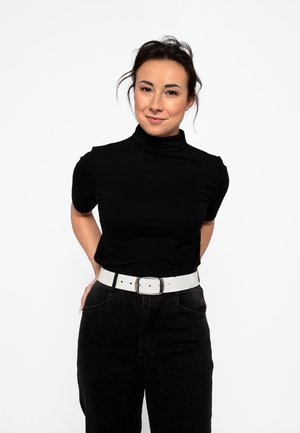 Ung kvinde med mørkt hår iført sort rullekrave og bukser, iført hvidt bælte, der står med hænderne bag ryggen og smiler mod en hvid baggrund.