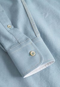 Primo piano di un tessuto di camicia azzurra chiara, con un polsino abbottonato dai dettagli bianchi, cuciture in contrasto e una texture liscia.