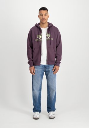 Purple zip-up hoodie with drawstrings, featuring a logo in light green. Worn over a white t-shirt and paired with blue jeans and sneakers.