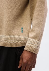 Μπεζ πλεκτό πουλόβερ με λευκά μανσέτα και τελείωμα με σχέδιο, διακοσμημένο με τρεις μικρές μπλε ραφές κοντά στη μέση, φορεμένο με μαύρο παντελόνι.