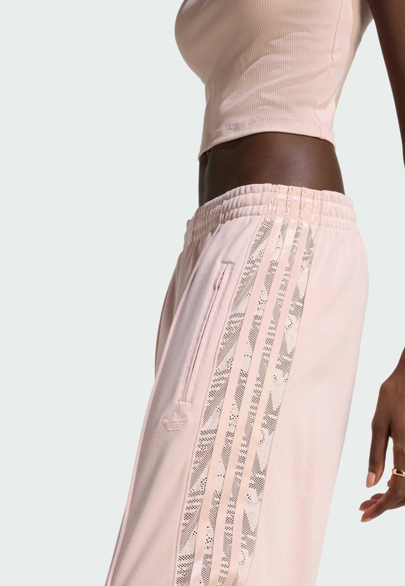 Light pink sweatpants with a textured waistband, side zipper pocket, and decorative side stripes featuring a lace pattern and logo accent.