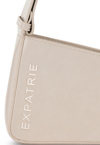 Borsa a mano in pelle beige chiaro con una texture liscia, forma angolare e logo "EXPATRIE" impresso sul lato. Dettaglio della tracolla regolabile visibile.