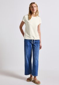 Haut léger crème texturé à manches courtes avec un ourlet à cordon, associé à un jean en denim bleu à jambe large et des sandales à enfiler en osier beige.