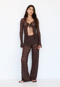 Rjava čipkast dvodelni komplet, ki vključuje dolg rokav cropped top z detajlom za zavezovanje spredaj in široke hlače s podobno čipkastim vzorcem.