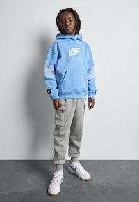 Hoodie Nike azul claro com logótipo branco, calças de moletom cinzas com bolsos e sapatos brancos com detalhes azuis. O fundo é simples.