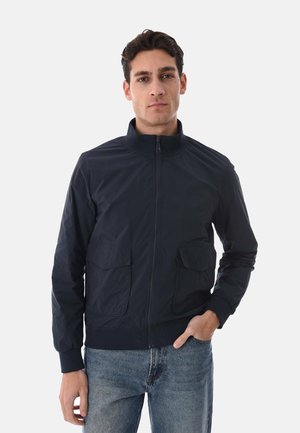Uomo che indossa una giacca blu navy con cerniera e due tasche frontali con patta, e jeans blu, in piedi con una mano in tasca su sfondo semplice.