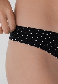 Černý bikiny s puntíky a hladkým povrchem, s širokým elastickým pasem. Pozadí ukazuje neutrální tón pleti.