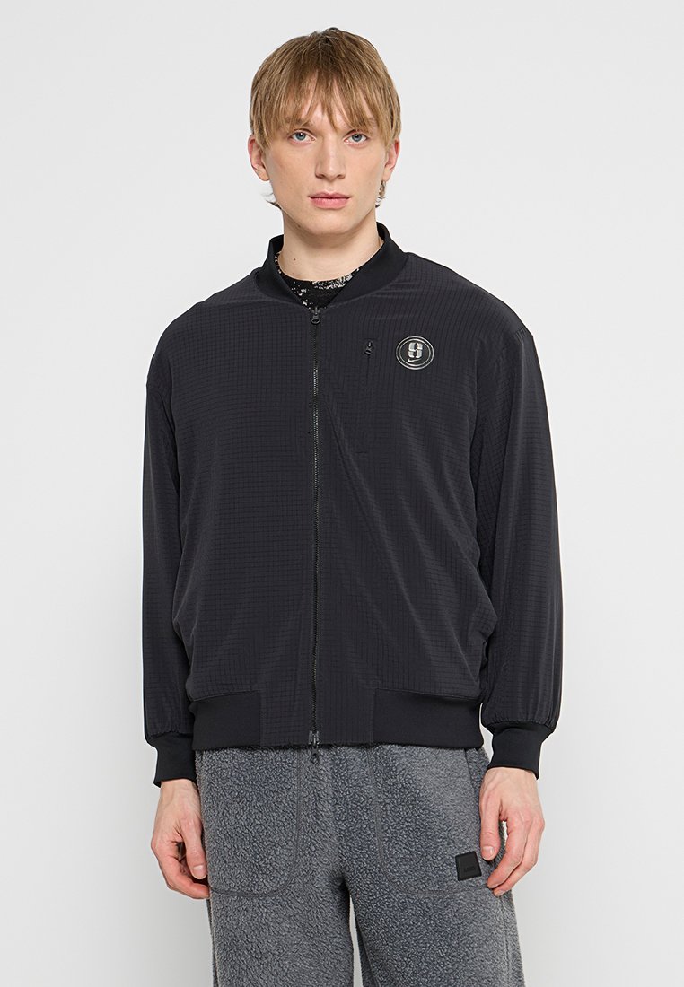 Schwarze Zip-Jacke aus strukturiertem Stoff, mit Stehkragen, elastischen Bündchen und kleinem Logo auf der linken Brust.