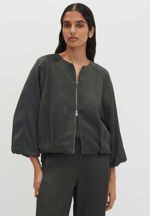 Donna con capelli lunghi e scuri che indossa una giacca nera ampia con zip e maniche a palloncino e pantaloni neri abbinati, in piedi davanti a uno sfondo chiaro e semplice.