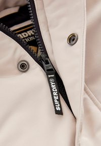 Jachetă bej deschis cu un fermoar negru, accesorii argintii și un etichetă de marcă pronunțată. Țesătură netedă cu un ușor luciu.
