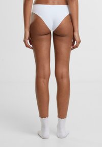 Fehér bikinialsó, magas szabású dizájnnal, minimális takarással és sima textúrával. A modellen fehér bokazokni van.