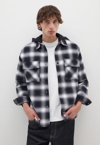 Schwarzes und weißes karriertes Flanellhemd mit zwei Brusttaschen, über einem weißen Hemd getragen, mit einer schwarzen Kapuze und Knopfverschluss.