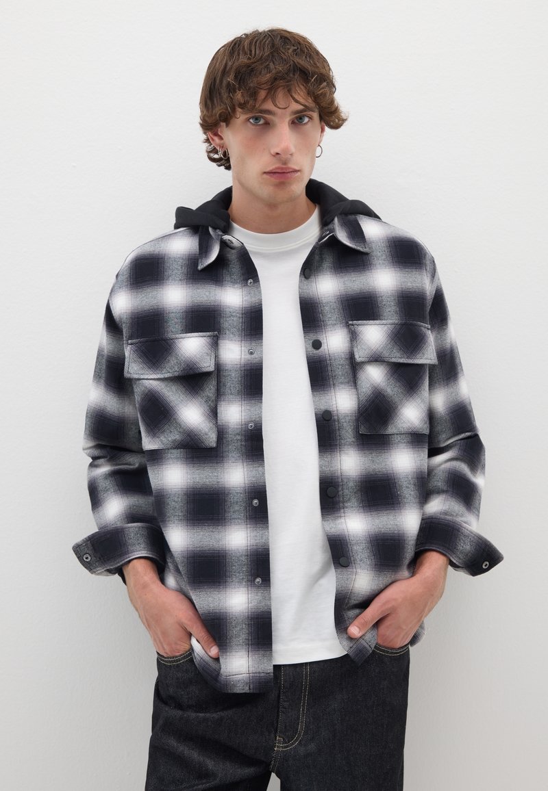 Schwarzes und weißes karriertes Flanellhemd mit zwei Brusttaschen, über einem weißen Hemd getragen, mit einer schwarzen Kapuze und Knopfverschluss.