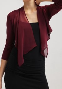 Burgundský průsvitný kardigan s tříčtvrtečními rukávy, otevřený vpředu a s volným střihem, vrstvený přes černé přiléhavé šaty. Jemná textura.