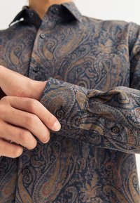 Meeste tumesinise nurknööbiga särk kullakarva, roostepunase ja burgundia paisleimustriga. Omab struktureeritud kraet ja mansetti mustade nuppudega.