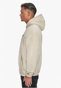 Beige hættetrøje i blødt stof, med en kængurulomme, ribbede manchetter og logo-detalje på brystet. Slim fit silhuet.