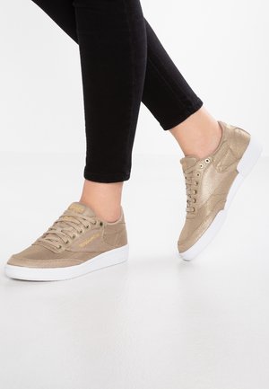 Nogi w czarnych spodniach noszące metaliczne złote sneakersy sznurowane z białymi podeszwami, stojące na gładkiej białej powierzchni.