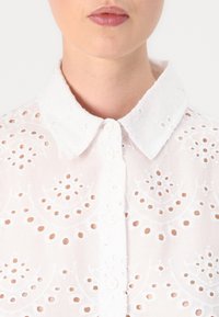 White embroidered blouse featuring a button-up front, a collared neck, and intricate cut-out designs with circular patterns throughout.