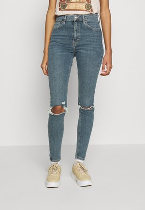 Kvinde iført blå skinny jeans med høj talje og knærevner, beige sneakers og en beige t-shirt med print mod en ensfarvet hvid baggrund.