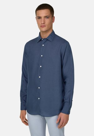 Mand iført en mørkeblå skjorte med lange ærmer og lysegrå bukser, stående mod en ensfarvet hvid baggrund.