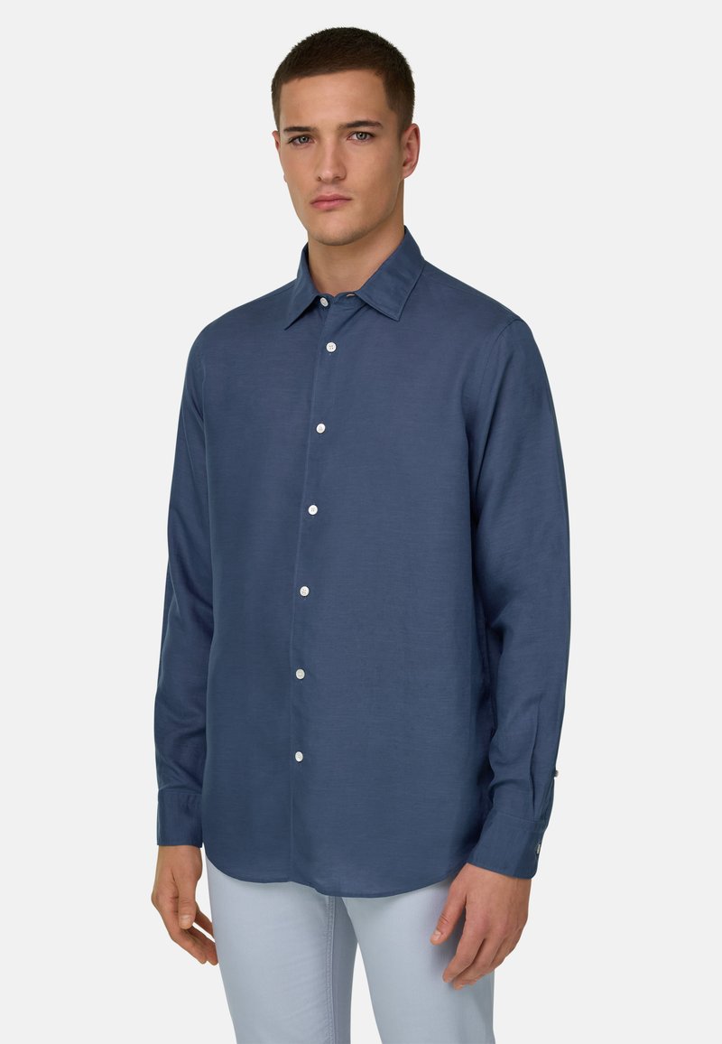 Homme portant une chemise bleu marine boutonnée à manches longues et un pantalon gris clair, debout devant un fond blanc uni.