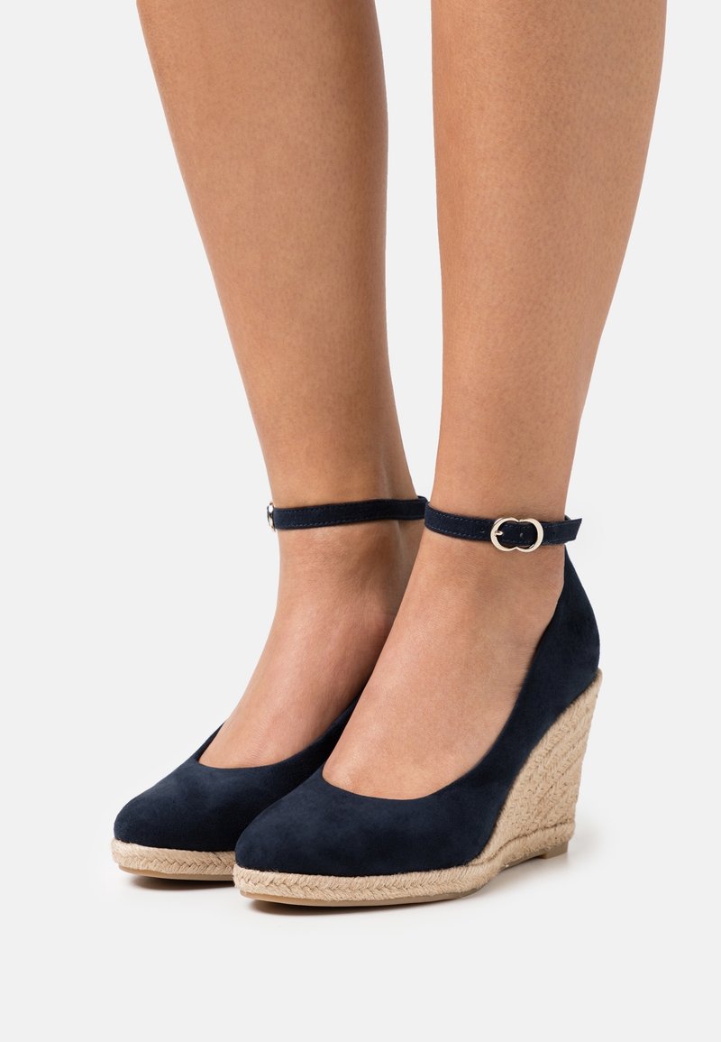 Jambes portant des compensées en daim bleu marine avec brides à la cheville et plateforme en jute tressée sur un fond clair uni.