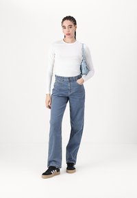 Denim hlače z črtami in sproščenim krojem, v kombinaciji z belim dolgim rokavom in črnimi supergami. Majhna modra torbica je nanešena čez ramo.