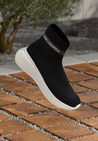 Sapatilha slip-on tricotada em preto com tornozelo alto, sola de borracha branca e logótipo "KARL LAGERFELD" na parte superior, com fundo em pedra e madeira.