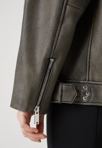 Grå lærjakke med teksturert finish, sølvglidelås, og justerbar stropp med metalliske detaljer ved mansjetten.