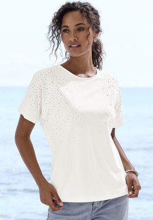 Woman with curly hair wearing a white short-sleeve shirt with small decorative dots, standing by the sea, hands in pockets.