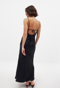 Black satin maxi dress with thin straps, an open back secured by ties, and a smooth, flowing texture. Length extends to the ankles.