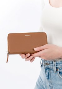 Hände halten eine braune Tamaris Lederbrieftasche mit Rundum-Reißverschluss vor weißem Oberteil und hellblauen Jeans.