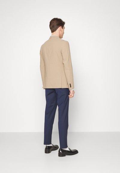 Blazer bege texturizado com um corte ajustado, combinado com calças azul-marinho. Mocassins pretos completam o visual. Detalhes de botões simples no blazer.