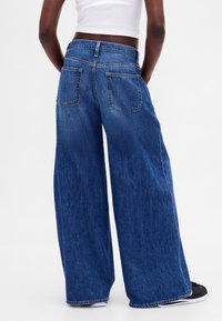 Riflě s širokými nohavicemi z modrého denimu, s vysokým pasem, dvěma zadními kapsami a třepeným lemem. Látka působí měkce a texturovaně.